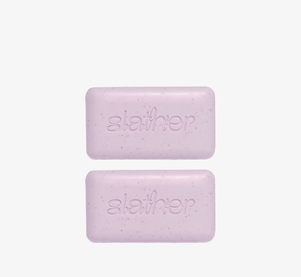 Exfoliating cleansing bars from Topicals Slather shown as a pair of speckled lavender bars with embossed branding on a white background.