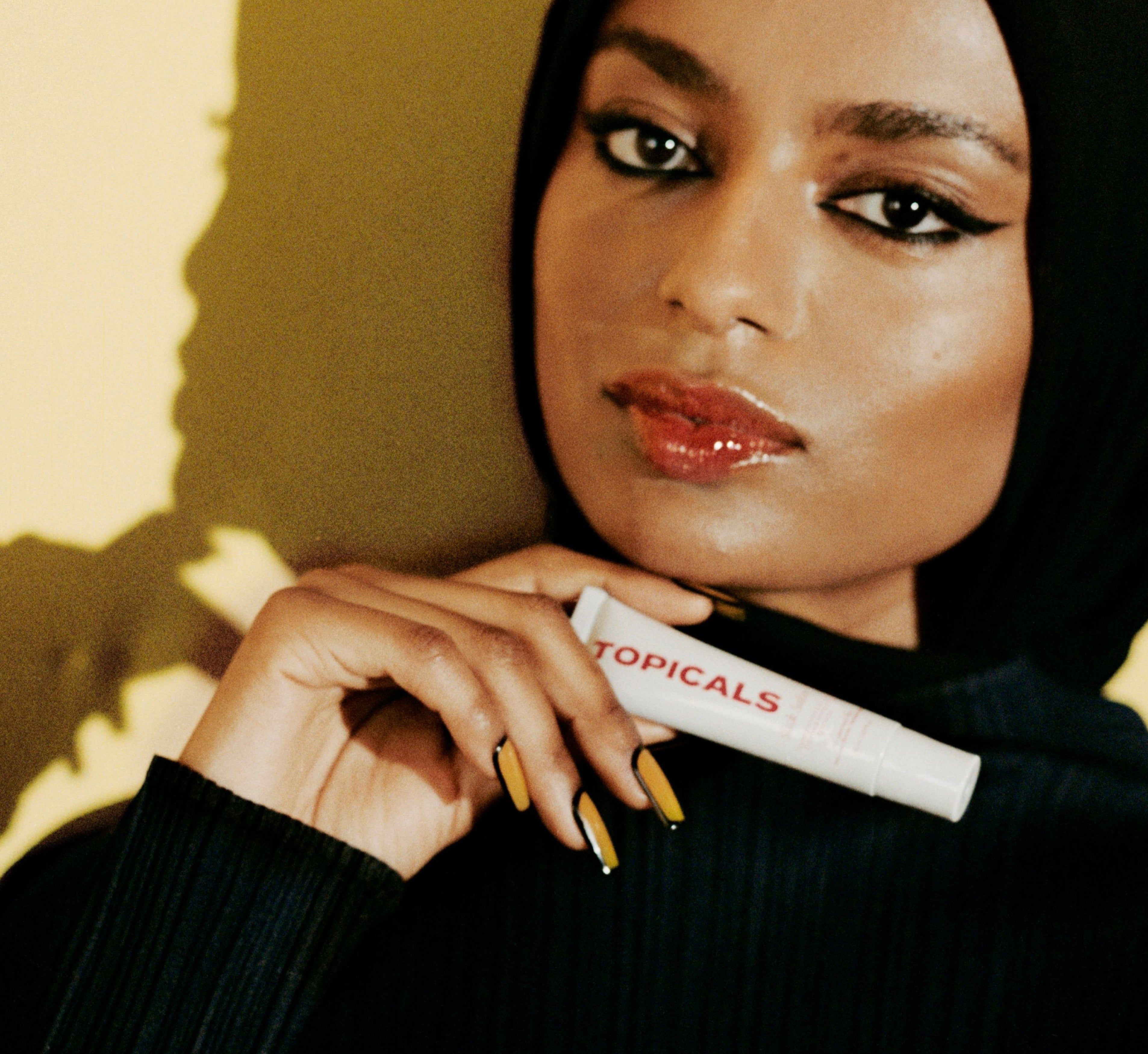 Moisturizing lip balm from Topicals Slick Salve held by a woman in a black hijab with glossy red lips and yellow nail art against a warm golden background.